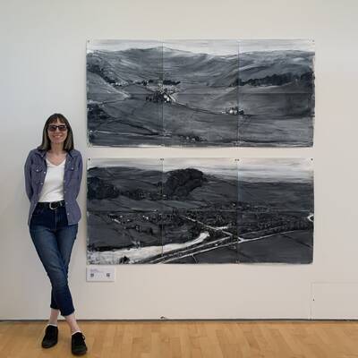 Deborah stands in front of her two large drawings in the gallery. she is wearing dark glasses and has bobbed brown hair. the drawings show the landscape as if from high up